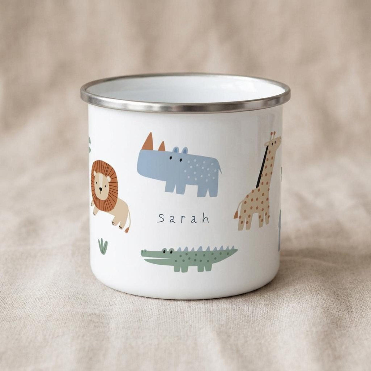 A white metal enamel mug with a silver rim displaying safari animal graphics and the name Sarah printed in the center, set against a neutral burlap background.