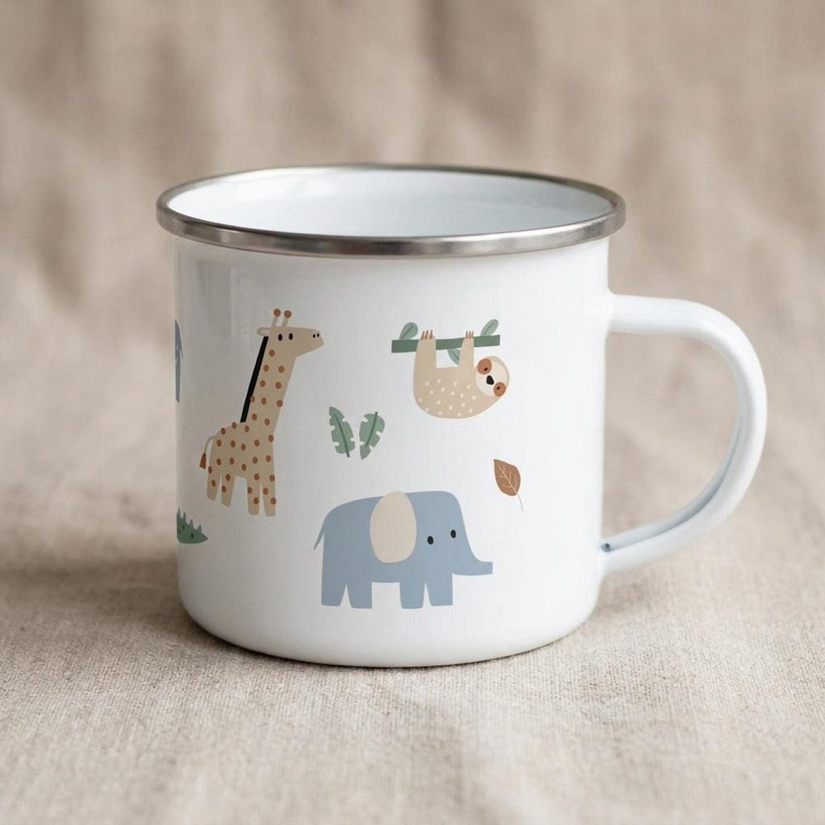 A white enamel camping mug featuring illustrations of a giraffe, elephant, and sloth among leaves, placed on a light beige fabric backdrop.