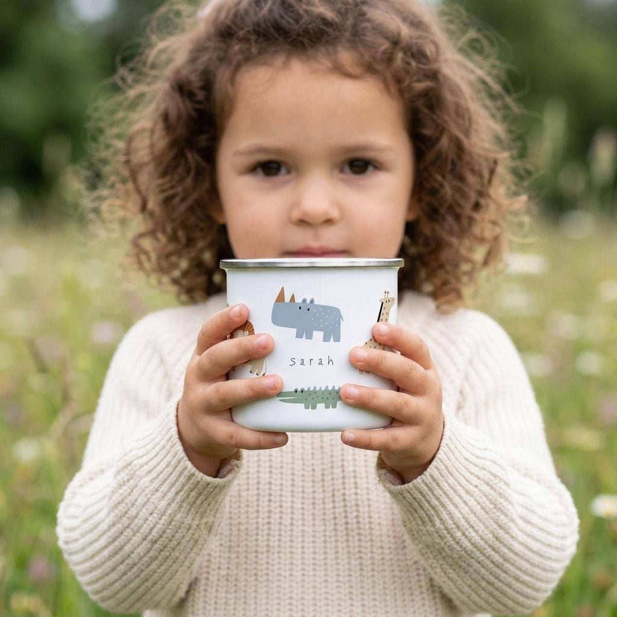 A young curly-haired child wearing a cream sweater holds a personalized safari-themed white enamel mug outdoors in a grassy field.