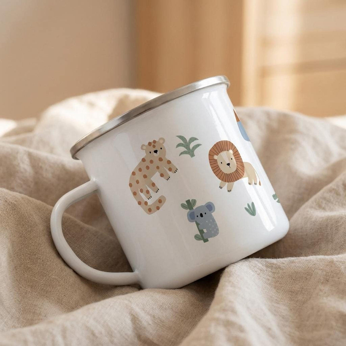 A white enamel mug with a silver rim decorated with cute safari animal drawings rests on a folded beige linen cloth.