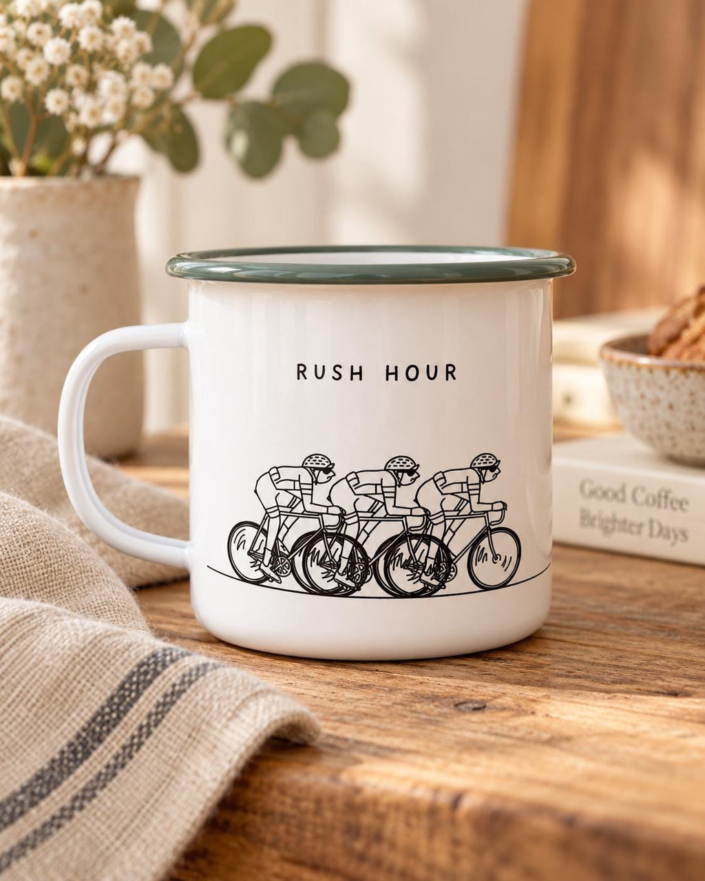 A white enamel mug with a dark green rim and a handle. The mug features a black line drawing of three cyclists riding in a row with the text RUSH HOUR printed above the illustration in a simple, clean, black sans-serif font.
