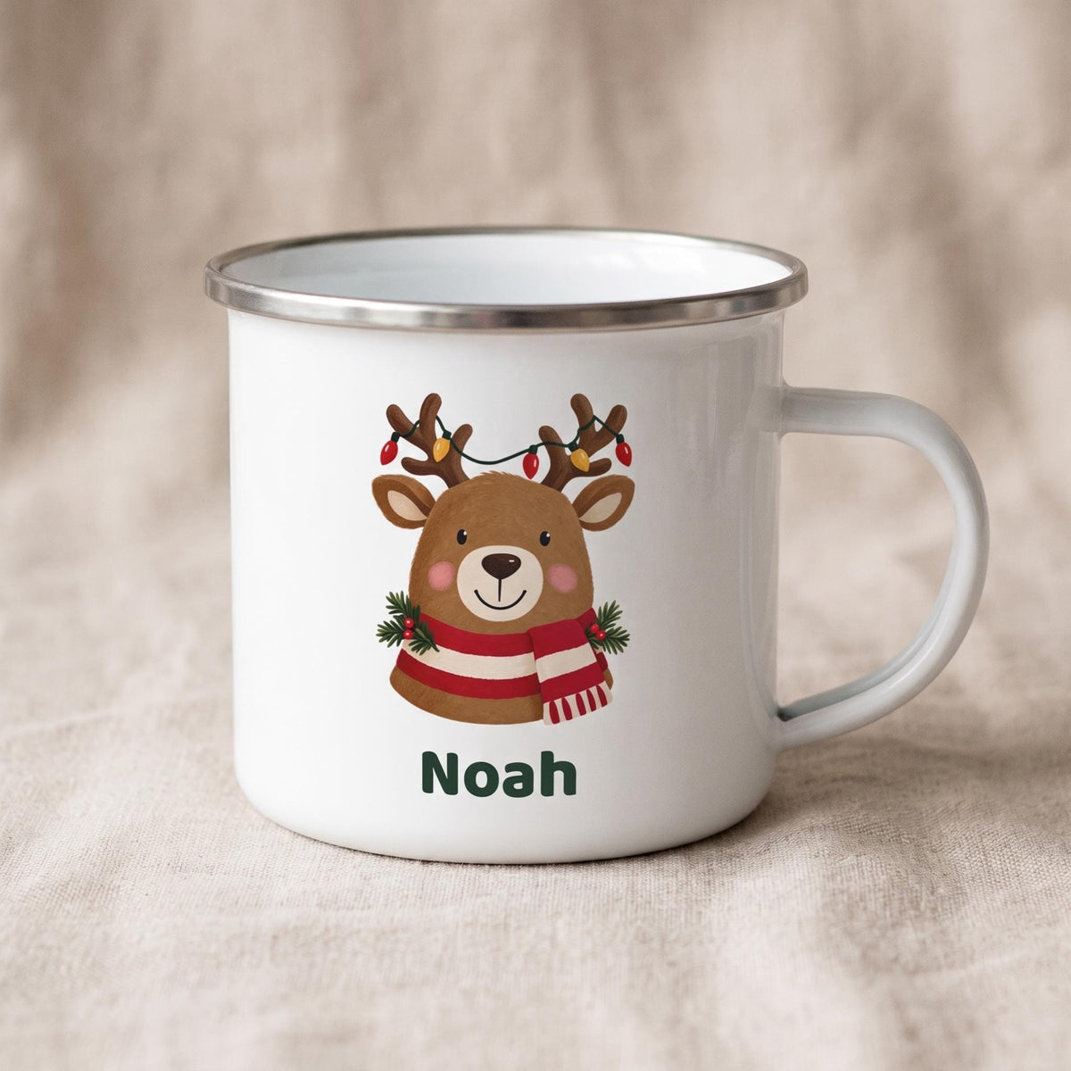 A white enamel mug with a silver metal rim displays a reindeer illustration with the name Noah on a beige linen background.