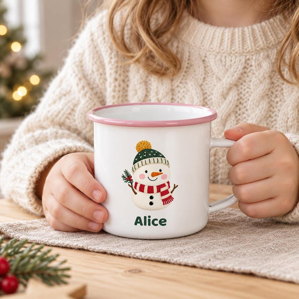 A child in a cream knit sweater holds a white enamel mug with a pink rim, featuring a snowman graphic and the name Alice.