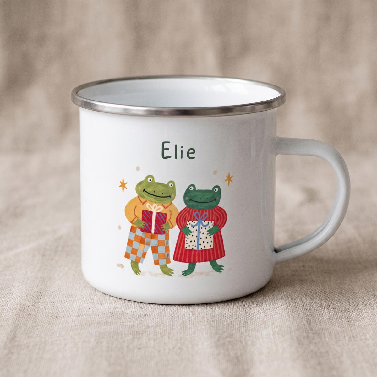 A white enamel mug with a silver rim featuring the name Elie and an illustration of two frogs holding wrapped presents.