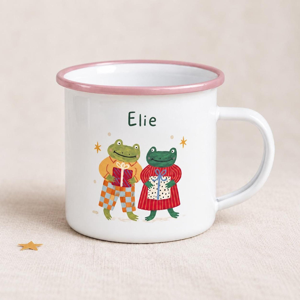 A white enamel mug with a pink rim showing the name Elie and an illustration of two frogs holding wrapped gifts.