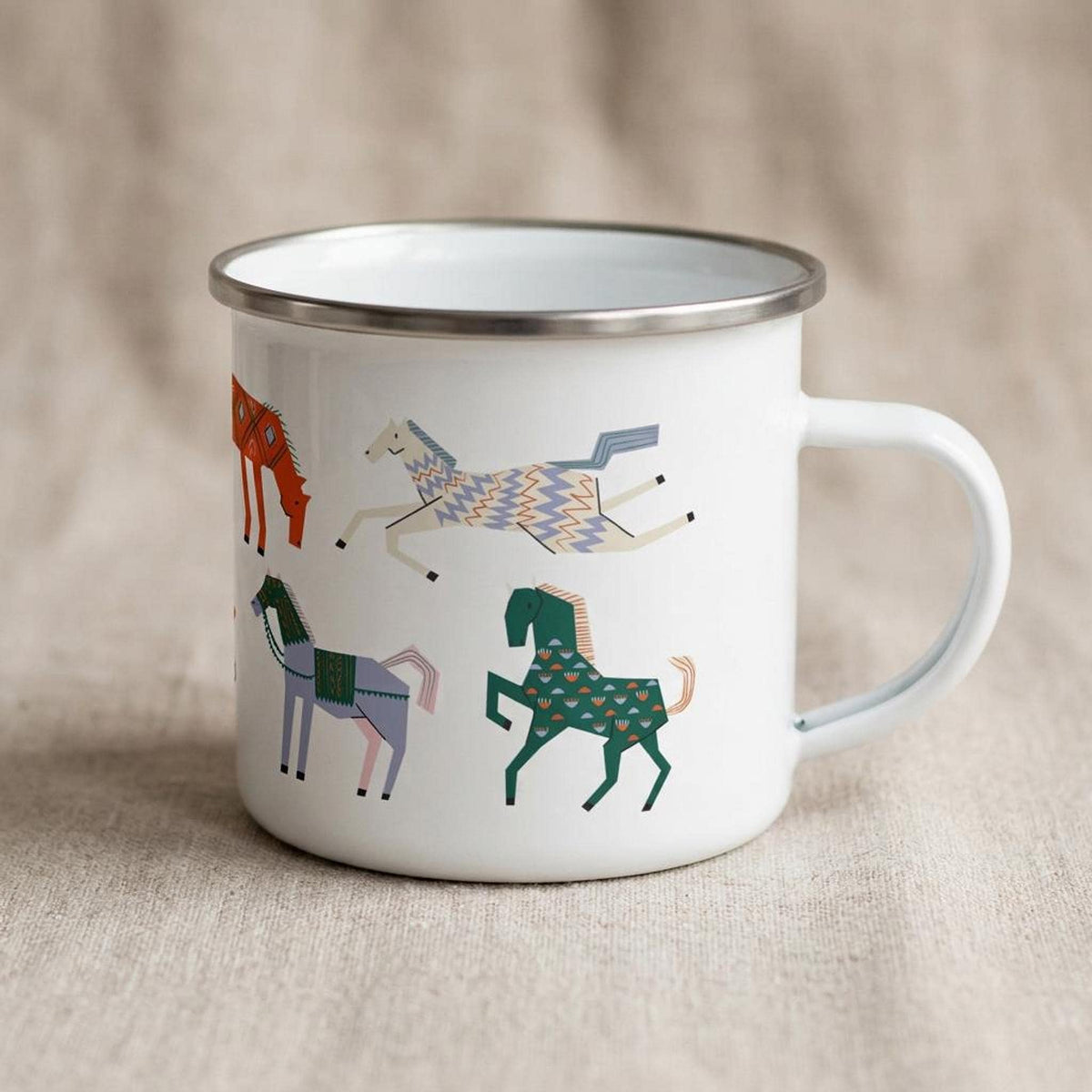 A white enamel mug featuring retro horse motifs is positioned on a light brown cloth background.