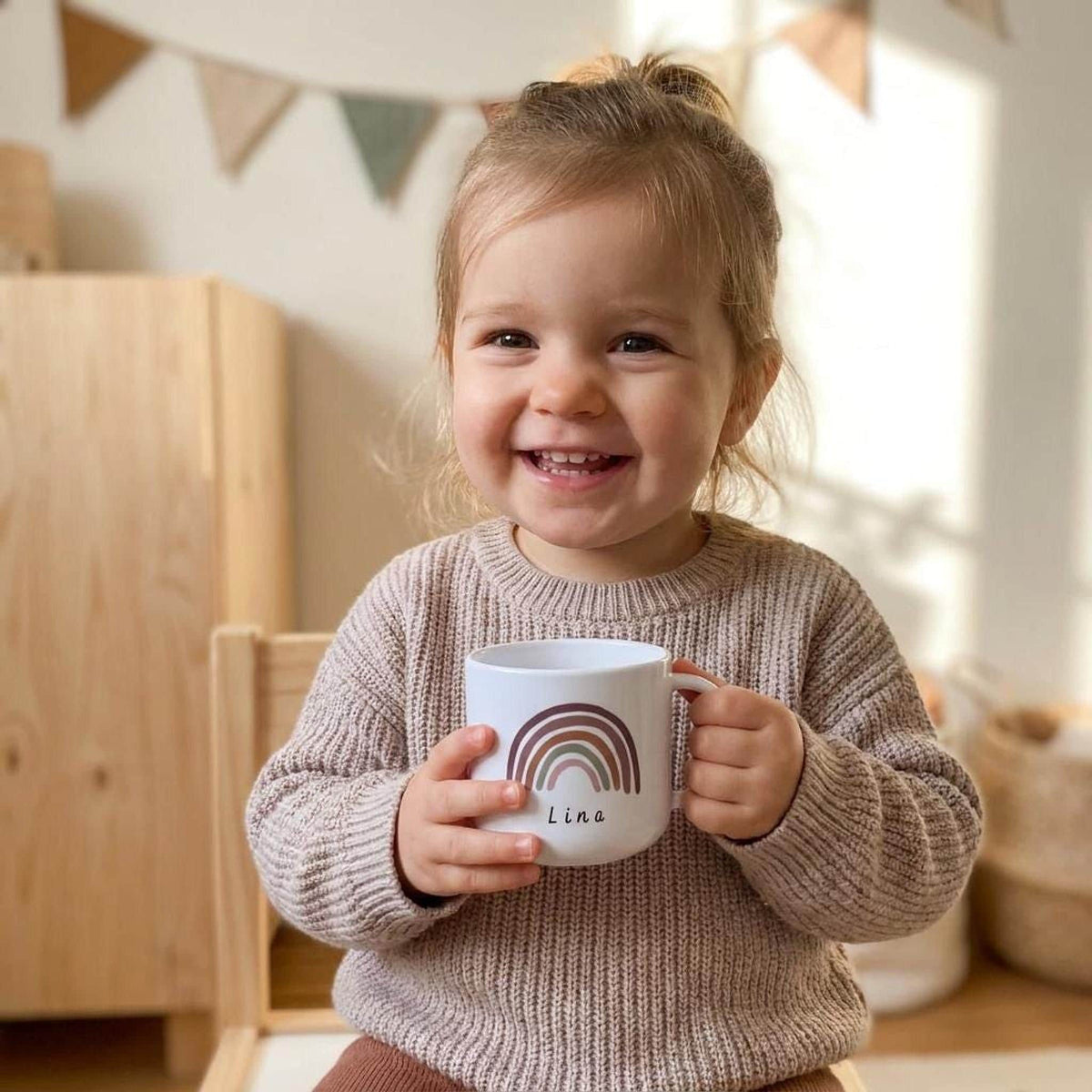 A smiling toddler wearing a beige sweater holds a white mug with a warm-toned rainbow and the name Lina.