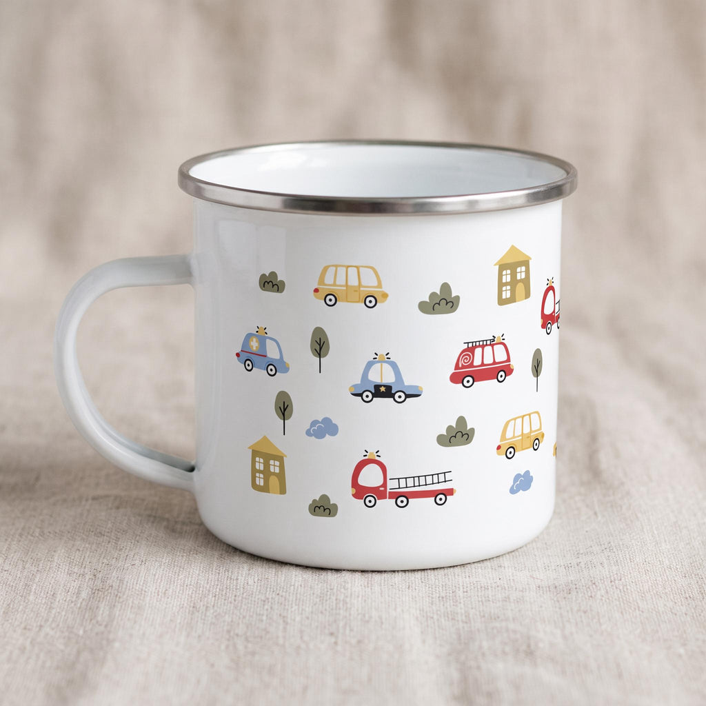 A white enamel mug with a silver rim and handle, decorated with cartoon fire trucks, police cars, ambulances, and houses against a neutral backdrop.