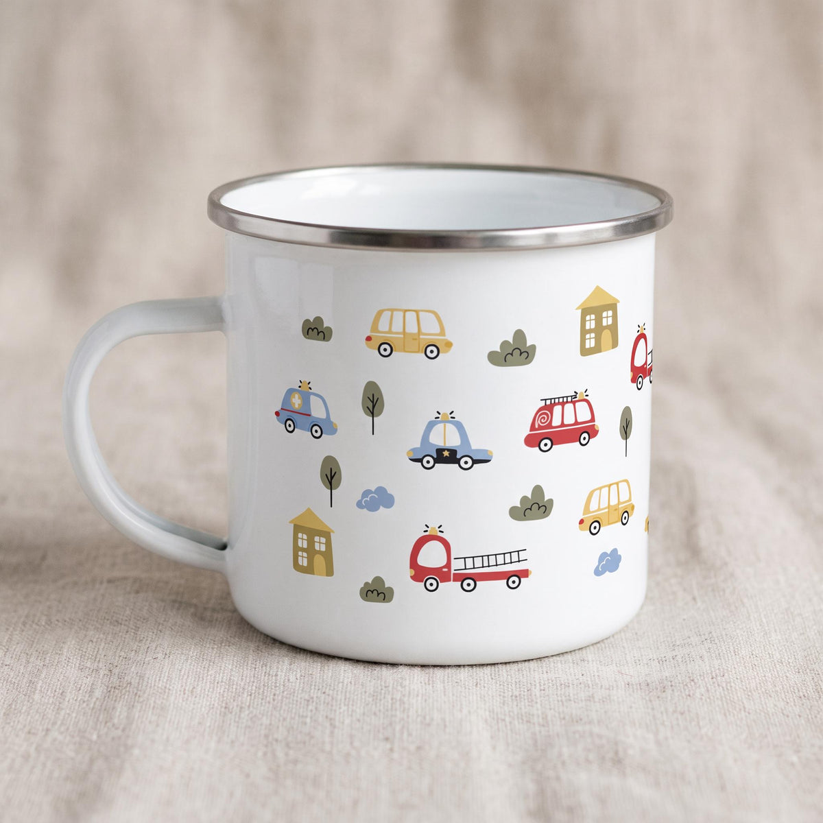 A white enamel mug with a silver rim and handle, decorated with cartoon fire trucks, police cars, ambulances, and houses against a neutral backdrop.