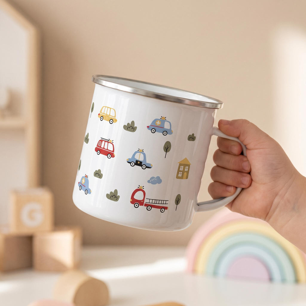 A hand holds a white enamel mug with a vehicle print against a neutral background featuring wooden blocks and a rainbow toy.