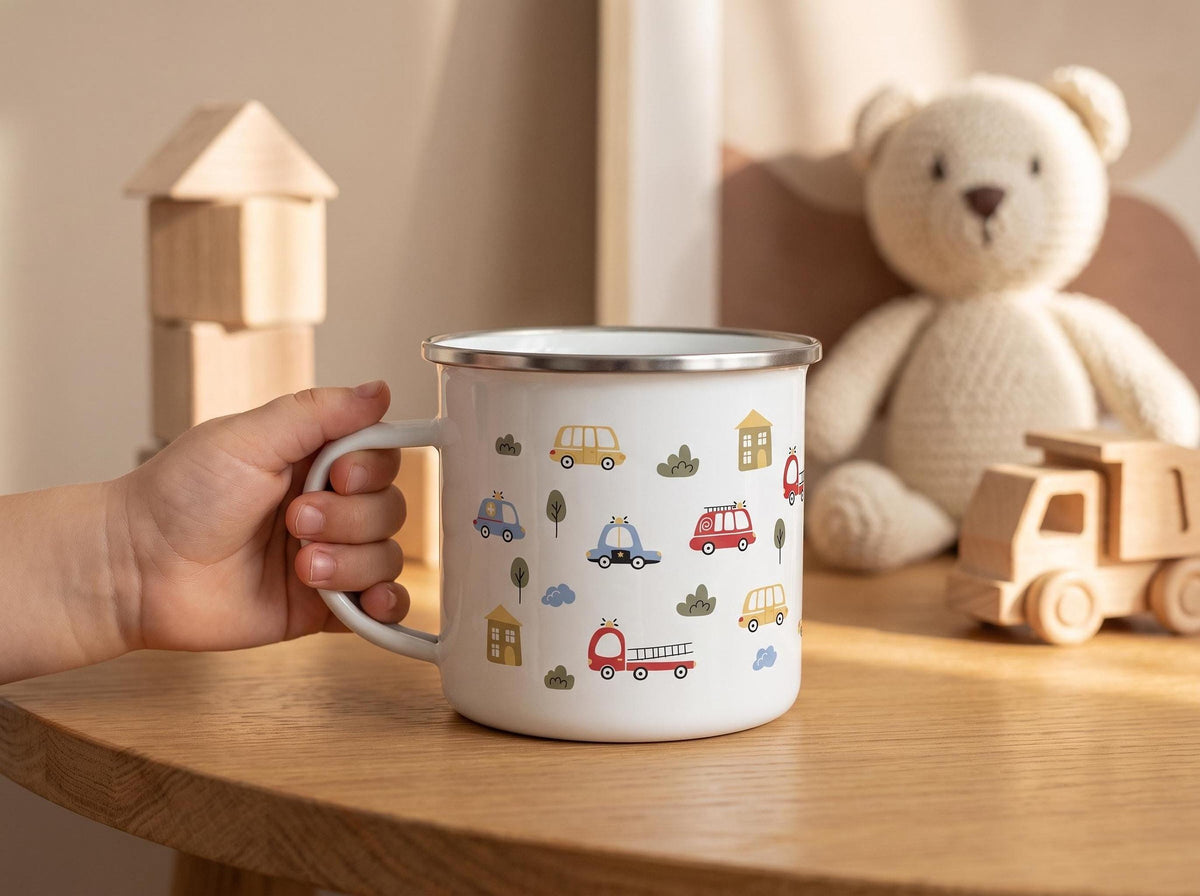 A child's hand holds a white enamel mug with a vehicle pattern on a wooden table, with toys in the blurred background.