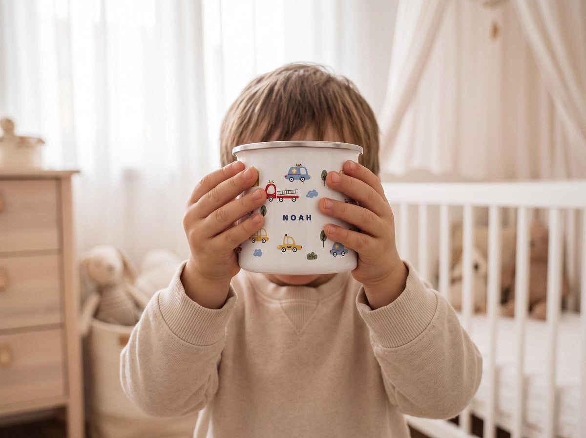 A child holds a white enamel mug featuring a name and vehicle illustrations in front of their face in a nursery room.