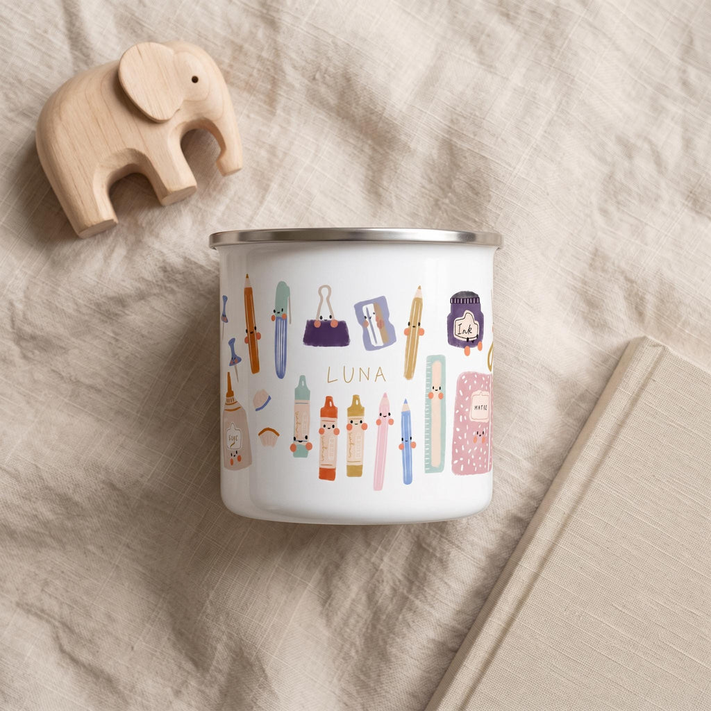 A personalised kids enamel mug featuring cute cartoon stationery and the name Luna stands on a neutral linen surface next to a wooden elephant toy and a notebook.
