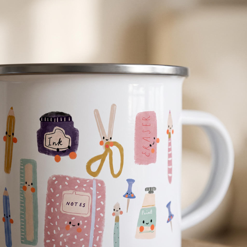 A close-up shot of a white enamel mug features detailed illustrations of cute stationery items with smiling faces, including an ink bottle, scissors, and a notebook.