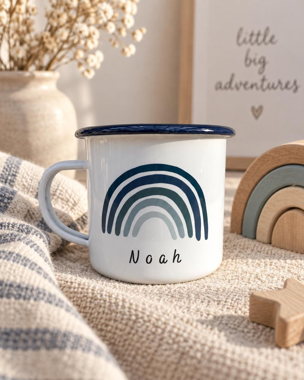 A white enamel mug with a dark blue rim and a handle. The front features a graphic of a multi-toned blue rainbow with the name Noah printed in black, lowercase, sans-serif lettering below the rainbow design.