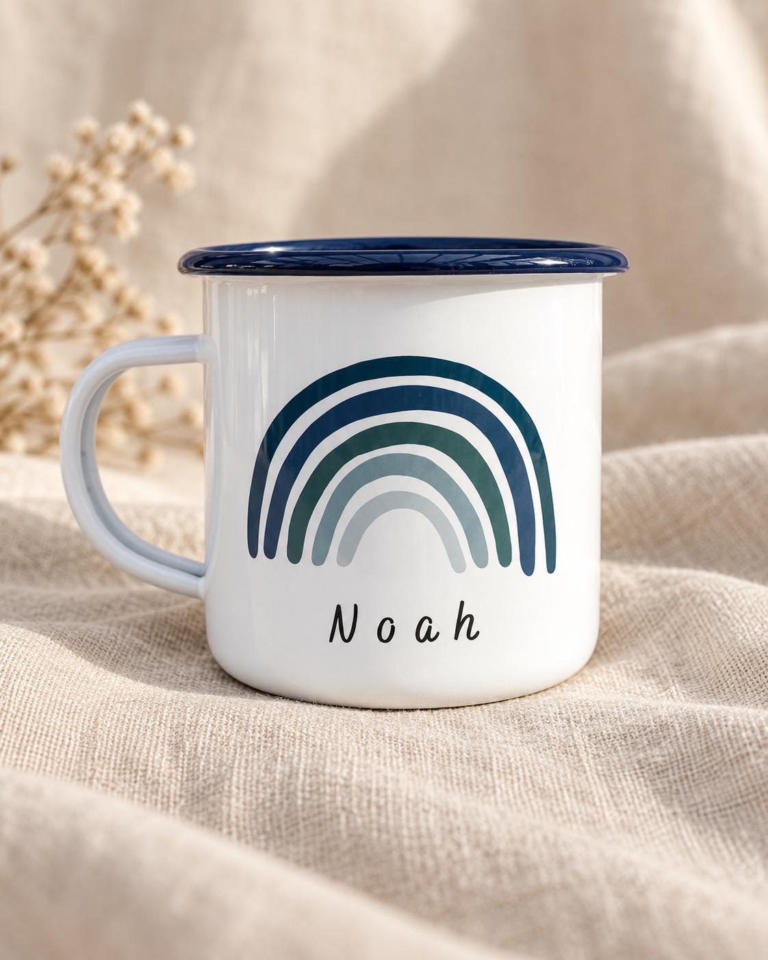 A white enamel mug with a dark blue rim and a handle. The front of the mug features a graphic of a rainbow in shades of blue and teal, with the name Noah printed in black, lowercase, sans-serif lettering centred below the rainbow.
