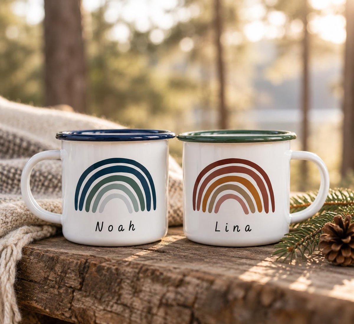 Two white enamel mugs with handles and coloured rims. The left mug has a dark blue rim and a blue-toned rainbow graphic with the name Noah. The right mug has a green rim and a warm-toned rainbow graphic with the name Lina.