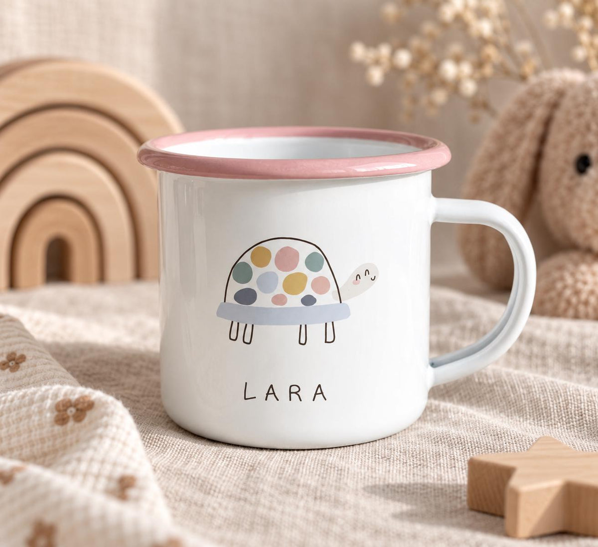 A white enamel mug with a pink rim and a handle, featuring a graphic of a turtle with a multi-coloured spotted shell and the name LARA printed in black capital letters below the turtle illustration on the front of the mug.