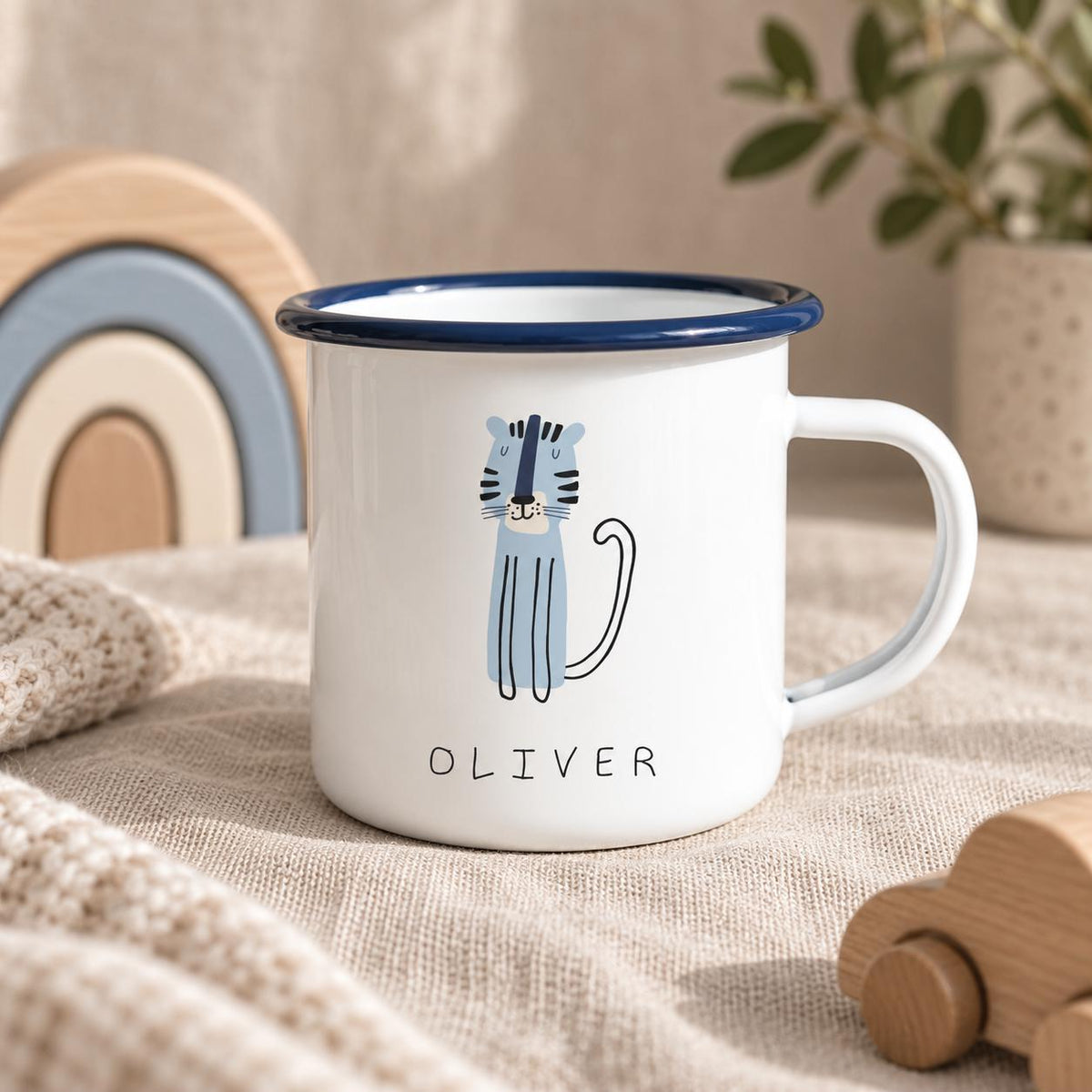 A white enamel mug with a dark blue rim and handle. The front features a light blue cartoon tiger illustration and the name OLIVER printed in black capital letters. The mug has a smooth, glossy finish and a simple cylindrical shape.