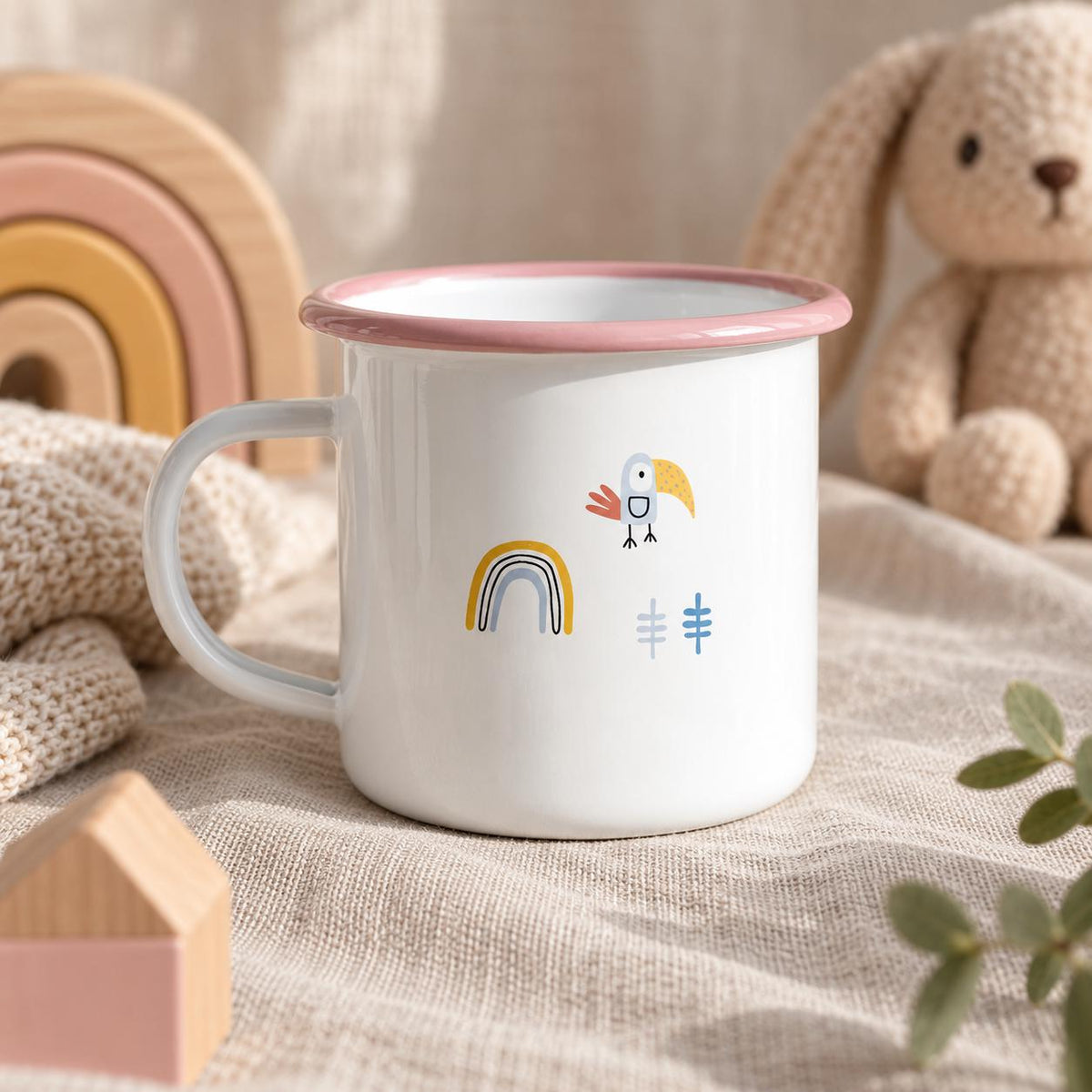 A white enamel mug with a pink rim and handle. The mug features a small graphic print consisting of a yellow and blue rainbow, a stylised bird with a yellow beak, and two small blue tree-like shapes on the front surface.