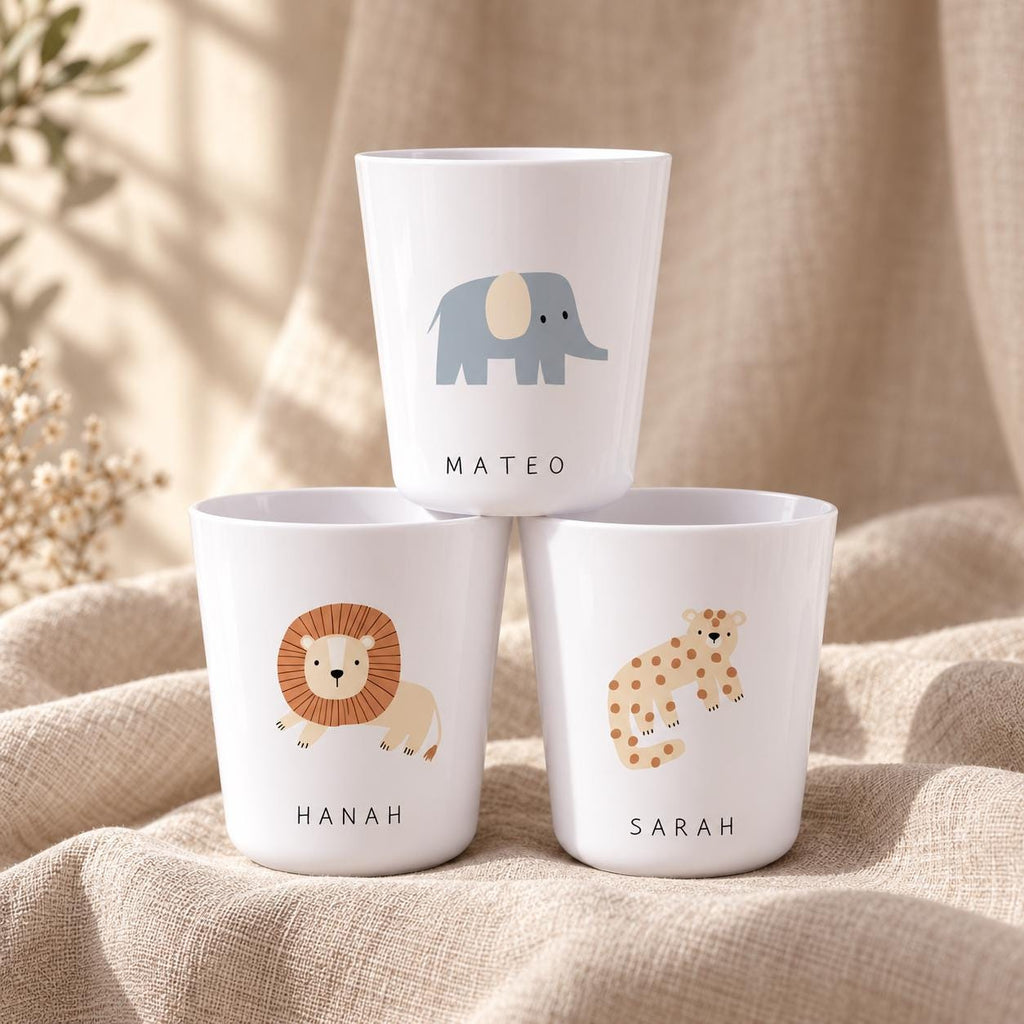 Three white plastic cups featuring animal illustrations and names. The top cup shows a grey elephant and the name MATEO. The bottom left cup features a brown lion and the name HANAH. The bottom right cup displays a spotted leopard and the name SARAH.