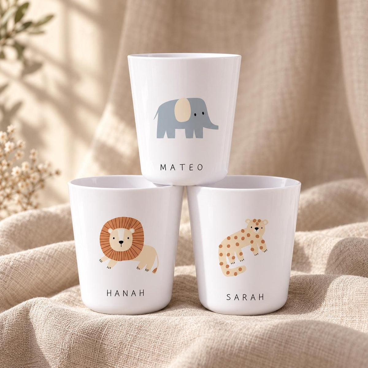 Three white plastic cups featuring animal illustrations and names. The top cup shows a grey elephant and the name MATEO. The bottom left cup features a brown lion and the name HANAH. The bottom right cup displays a spotted leopard and the name SARAH.