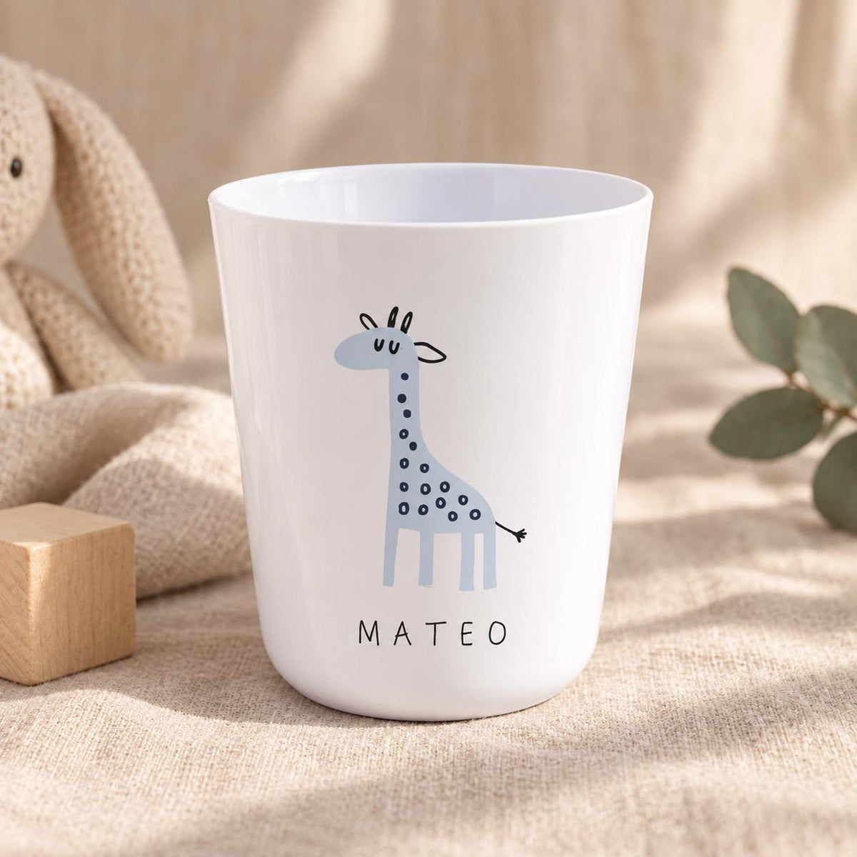 A white plastic cup featuring a light blue giraffe illustration with dark blue spots and the name MATEO printed in black capital letters below the giraffe. The cup has a smooth, tapered cylindrical shape with a wide, open top rim.
