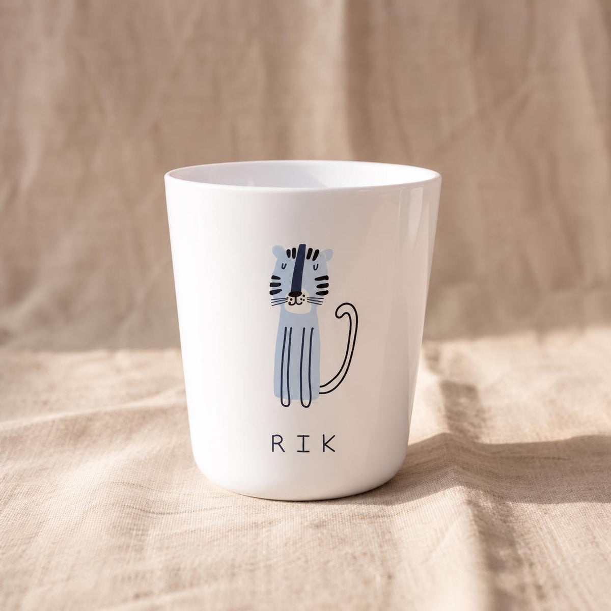 A white cylindrical cup featuring a light blue illustrated tiger with dark blue stripes and a long tail. Below the tiger, the name RIK is printed in simple black capital letters. The cup has a smooth finish and a tapered base.