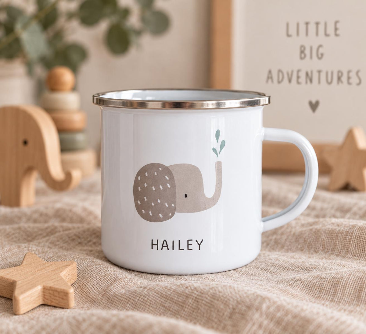 A white enamel mug with a silver rim, featuring a graphic of a light brown elephant with a small green water spray above its trunk. The name HAILEY is printed in black capital letters below the elephant illustration on the mug.