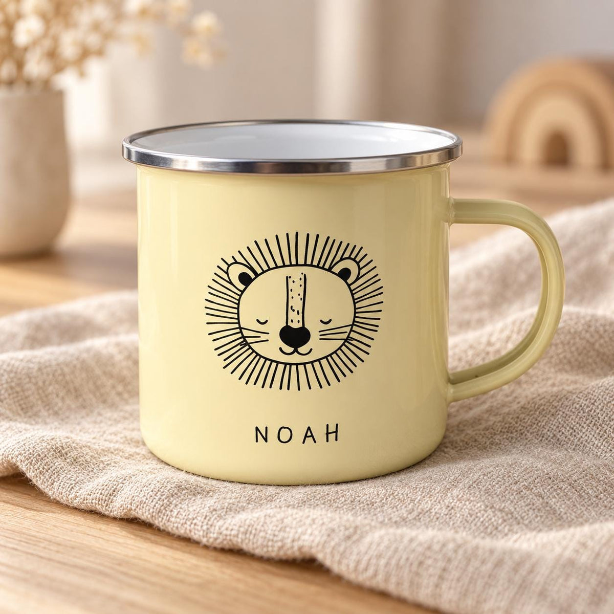 A pale yellow enamel mug featuring a black line drawing of a lion face with a mane and the name NOAH printed in black capital letters below the illustration. The mug has a smooth finish and a silver-coloured metal rim around the top edge.