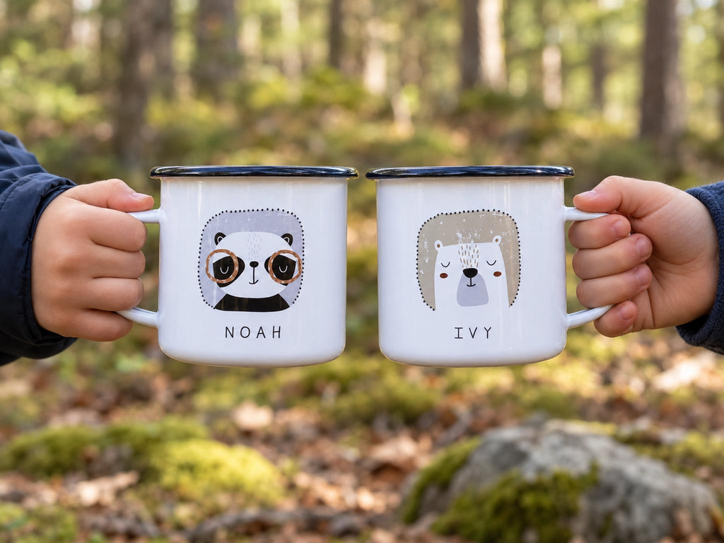 Two children's hands holding personalised panda and bear enamel mugs with the names NOAH and IVY in a sunlit forest.