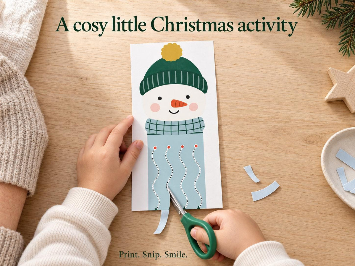 A child's hands use green scissors to cut along a snowman scarf activity at a wooden table, with a grown-up nearby.