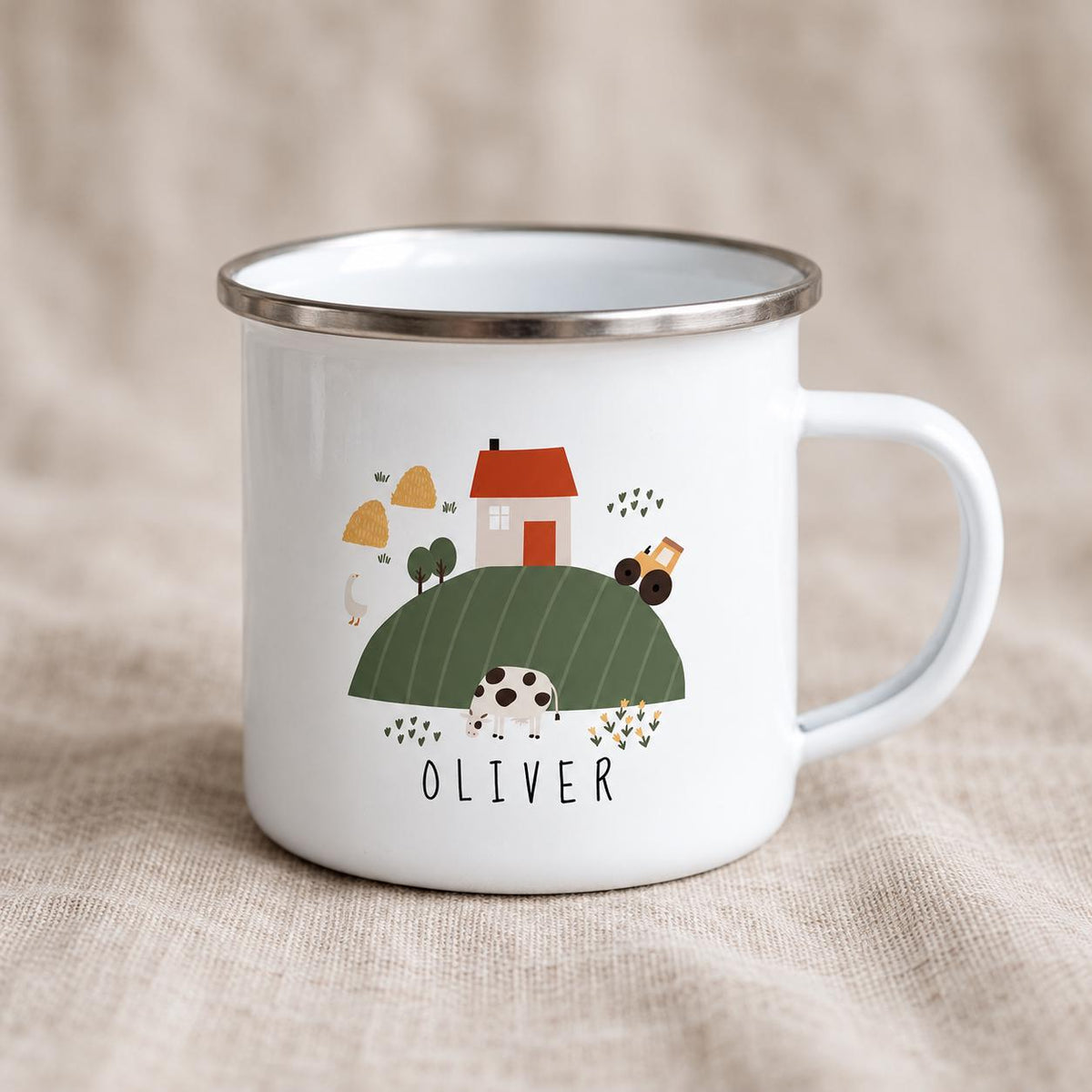 A white enamel mug with a silver rim and a handle, featuring a printed farm scene with a house, a cow, a tractor, and haystacks. The name OLIVER is printed in black capital letters below the green hill illustration on the mug.
