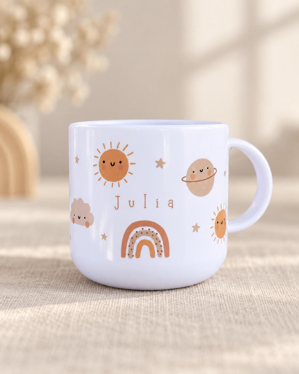 A white cup featuring a printed design of a smiling sun, a planet with rings, a cloud, and a rainbow. The name Julia is printed in the centre in brown letters. The cup has a rounded body and a side handle, with small stars scattered around.