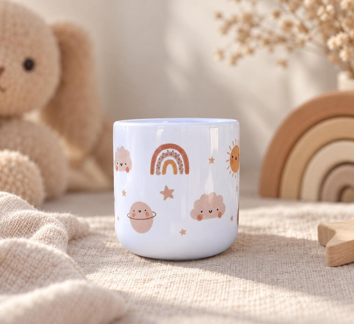 A white kids cup featuring a repeating pattern of small, smiling illustrations including a rainbow, a sun, clouds, stars, and a planet with rings in muted shades of brown, tan, and soft pink against a smooth, glossy surface.