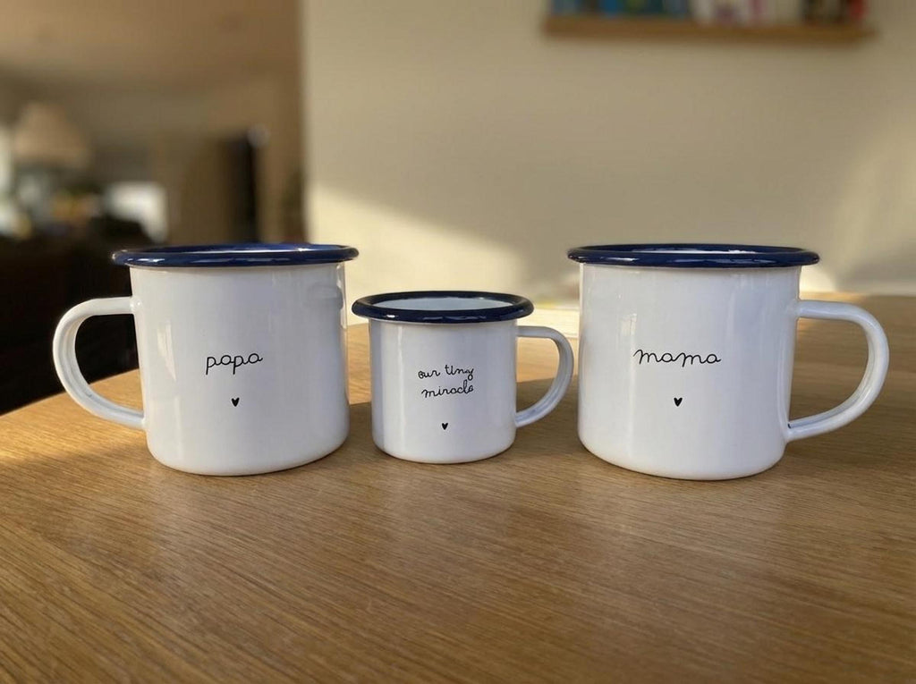 Three white enamel mugs with blue rims set on a wooden table with a blurred background showing papa, our tiny miracle, and mama.