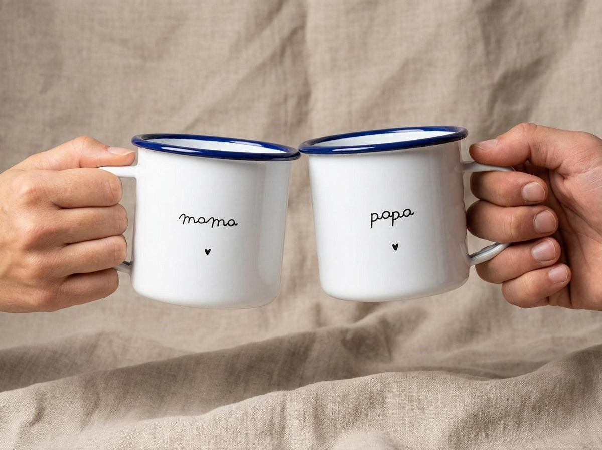 Two hands holding up a mama mug and a papa mug facing each other against a neutral fabric background.