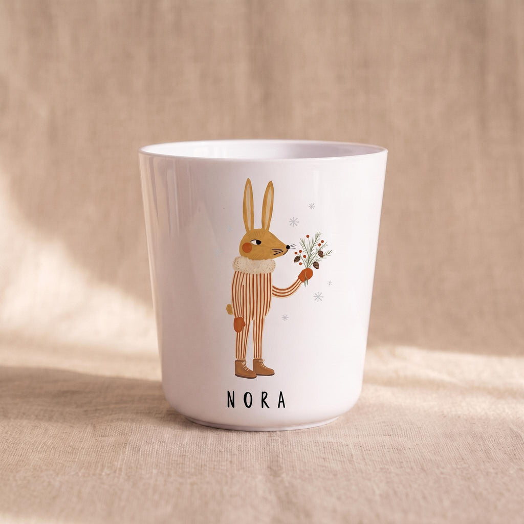 A single white child's cup featuring an illustration of a rabbit holding pine branches and the name Nora printed below, on a neutral fabric backdrop.