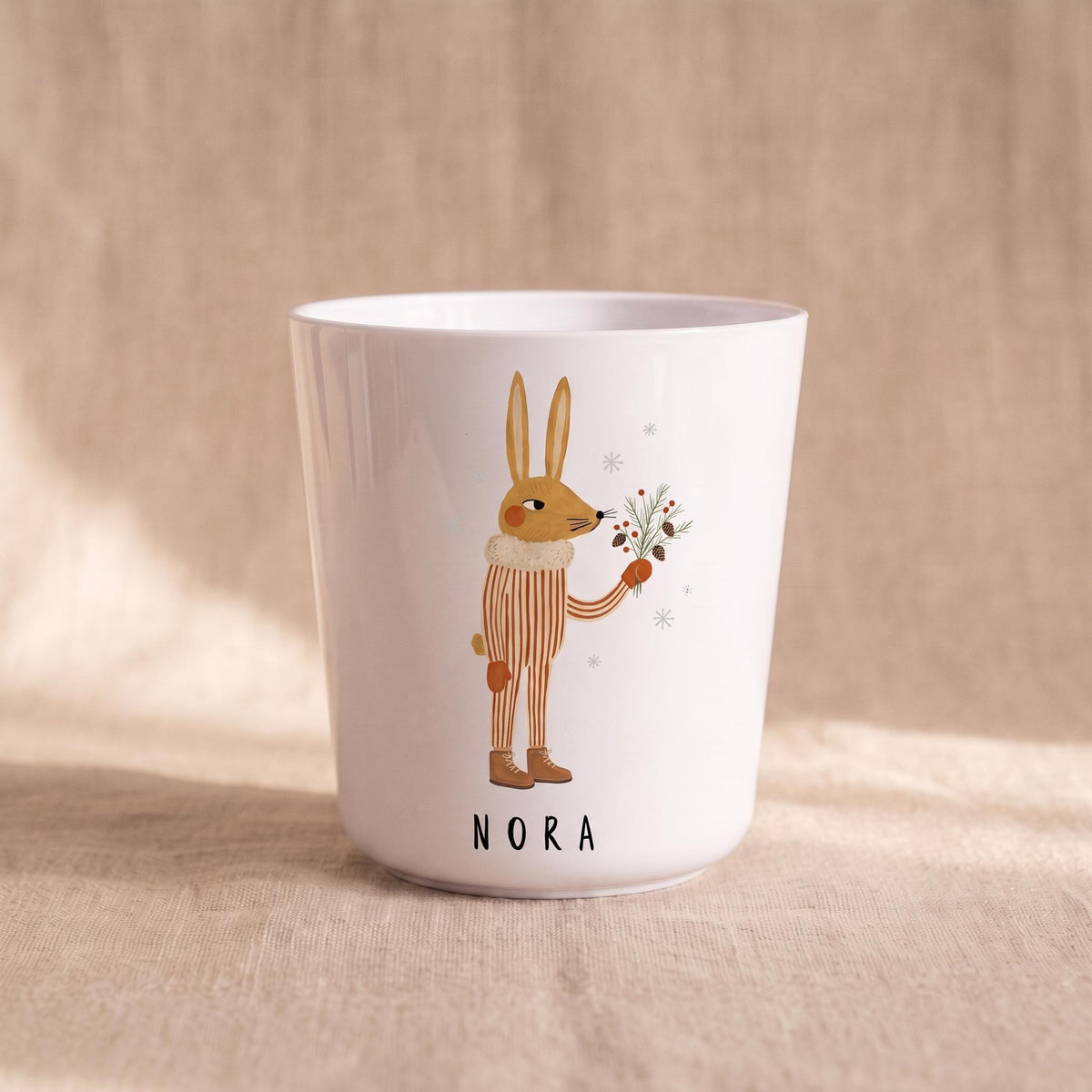 A single white child's cup featuring an illustration of a rabbit holding pine branches and the name Nora printed below, on a neutral fabric backdrop.
