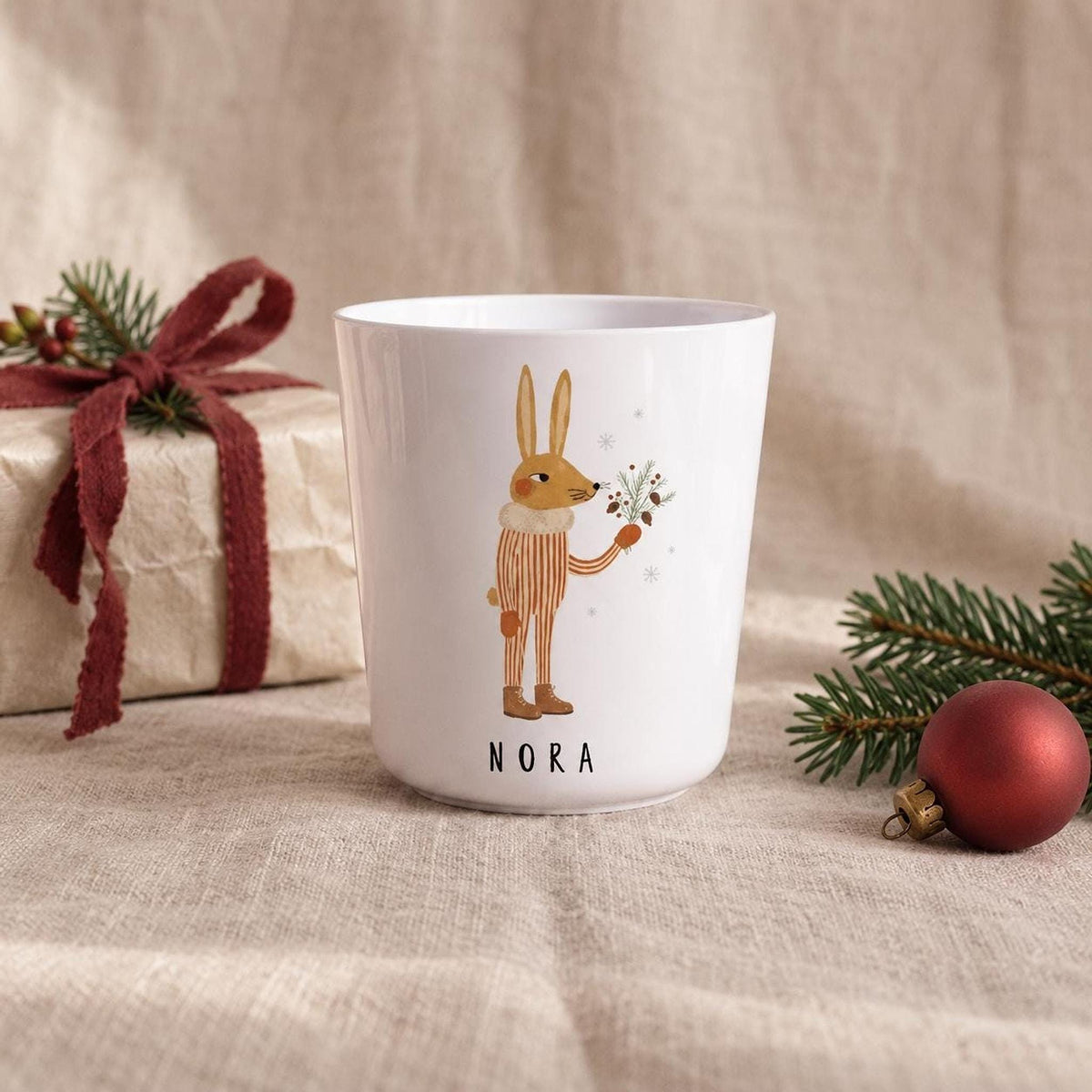 A white child's cup with an illustration of a rabbit holding pine sprigs and the name Nora printed below, set on a textured fabric cloth next to a wrapped gift and an ornament.