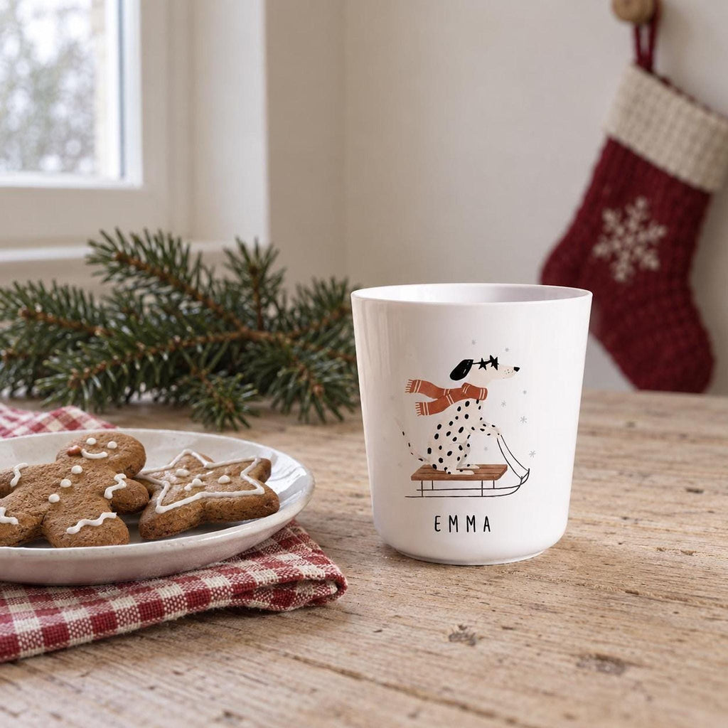 A white child's cup featuring an illustration of a dalmatian on a sled and the name Emma printed below, displayed on a wooden table next to a plate of gingerbread cookies.