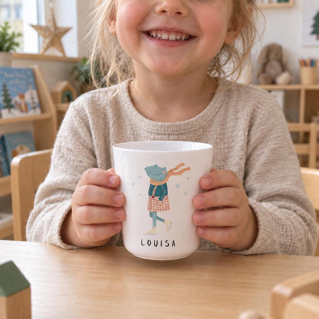 A smiling child holding a white cup with an illustration of an ice-skating frog and the name Louisa printed on it, sitting at a wooden table.