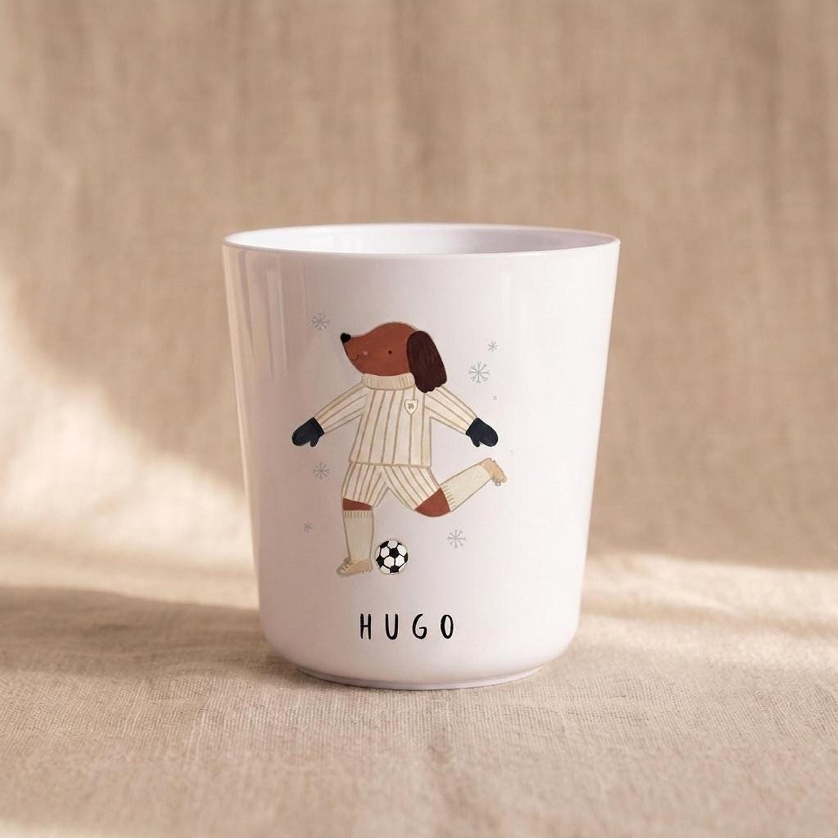 A single white child's cup featuring an illustration of a dog playing soccer and the name Hugo printed at the bottom, placed on a textured tan background.