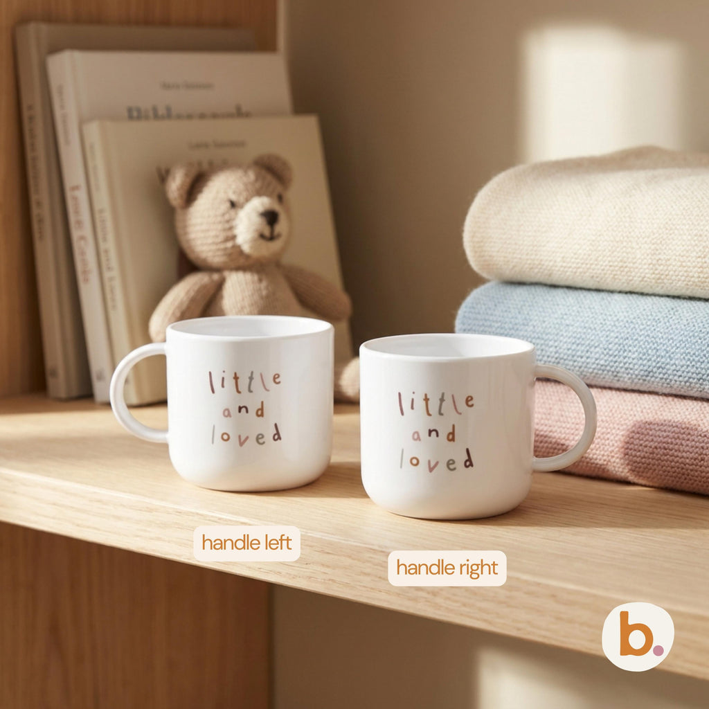 Two white mugs printed with little and loved on a wooden shelf next to stacked knit blankets and a toy bear.