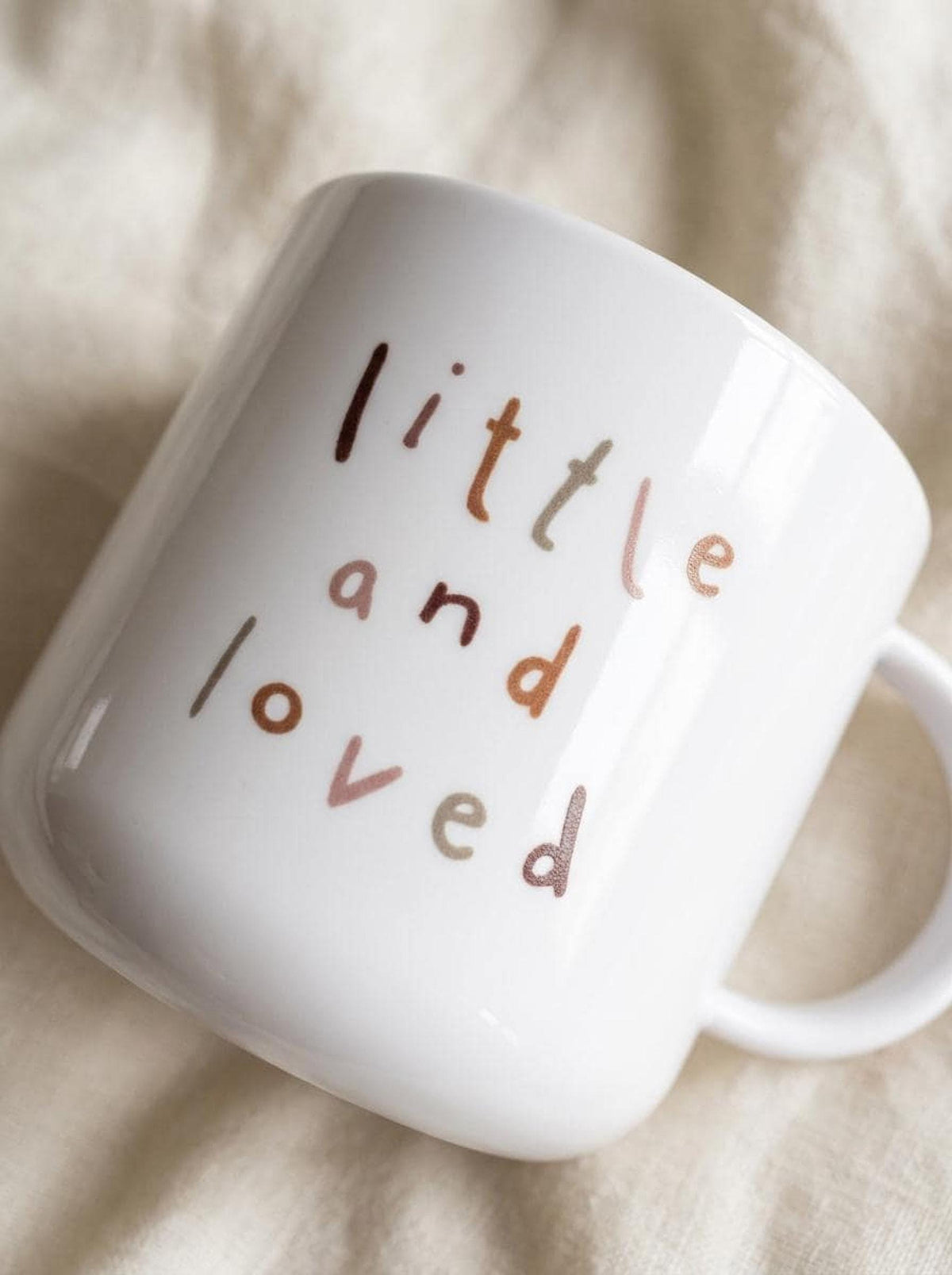 Close up view of a white mug printed with the words little and loved in brown lettering on a neutral fabric backdrop.