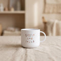 A white mug with little and loved printed on the front, sitting on a neutral tablecloth in a softly lit room.