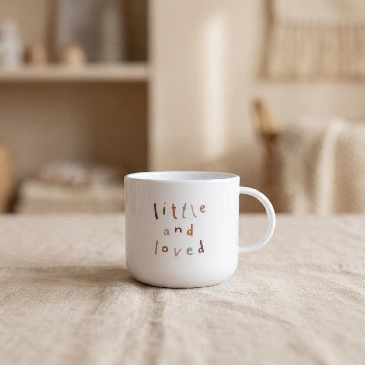 A white mug with little and loved printed on the front, sitting on a neutral tablecloth in a softly lit room.
