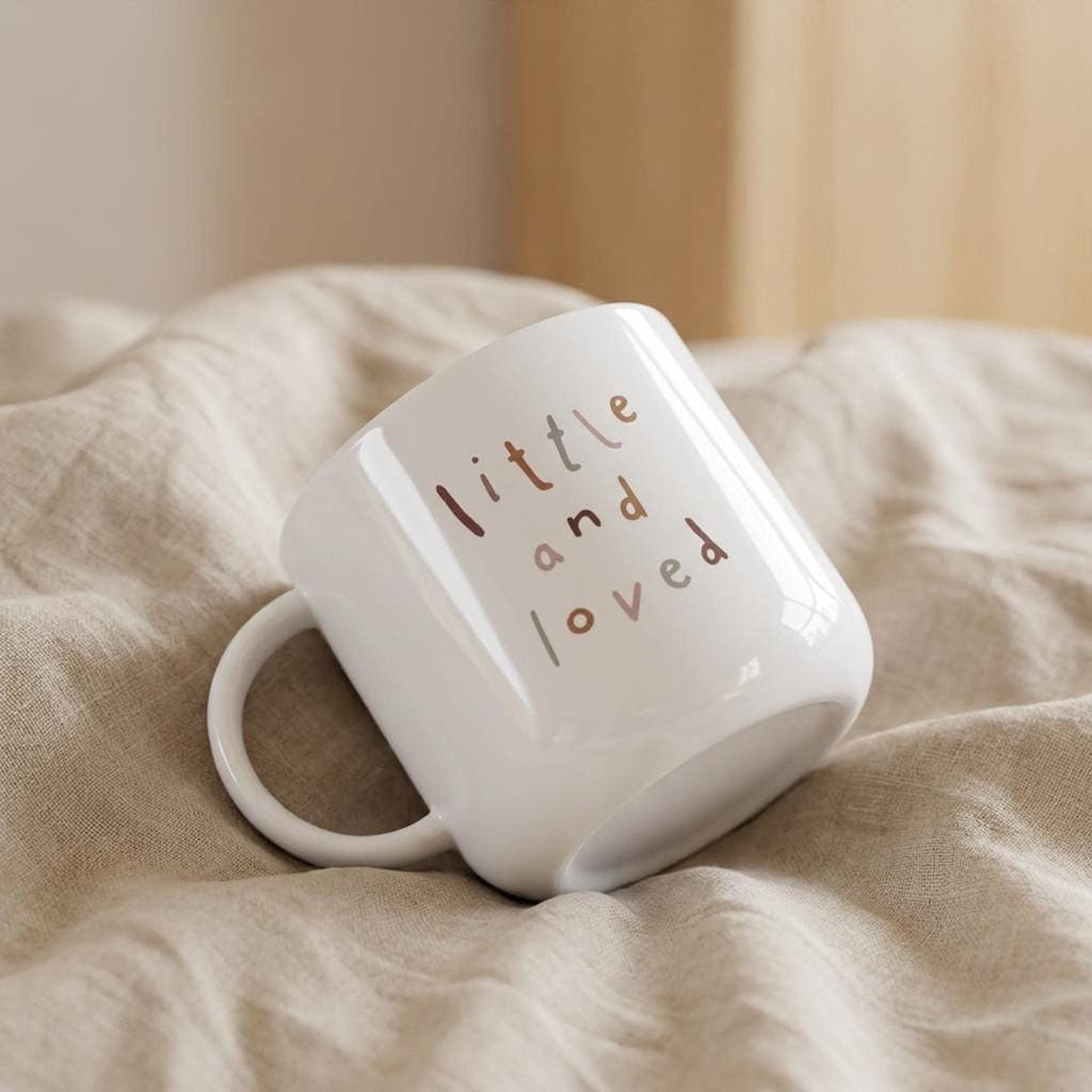 A white mug printed with little and loved lying sideways on wrinkled beige fabric.