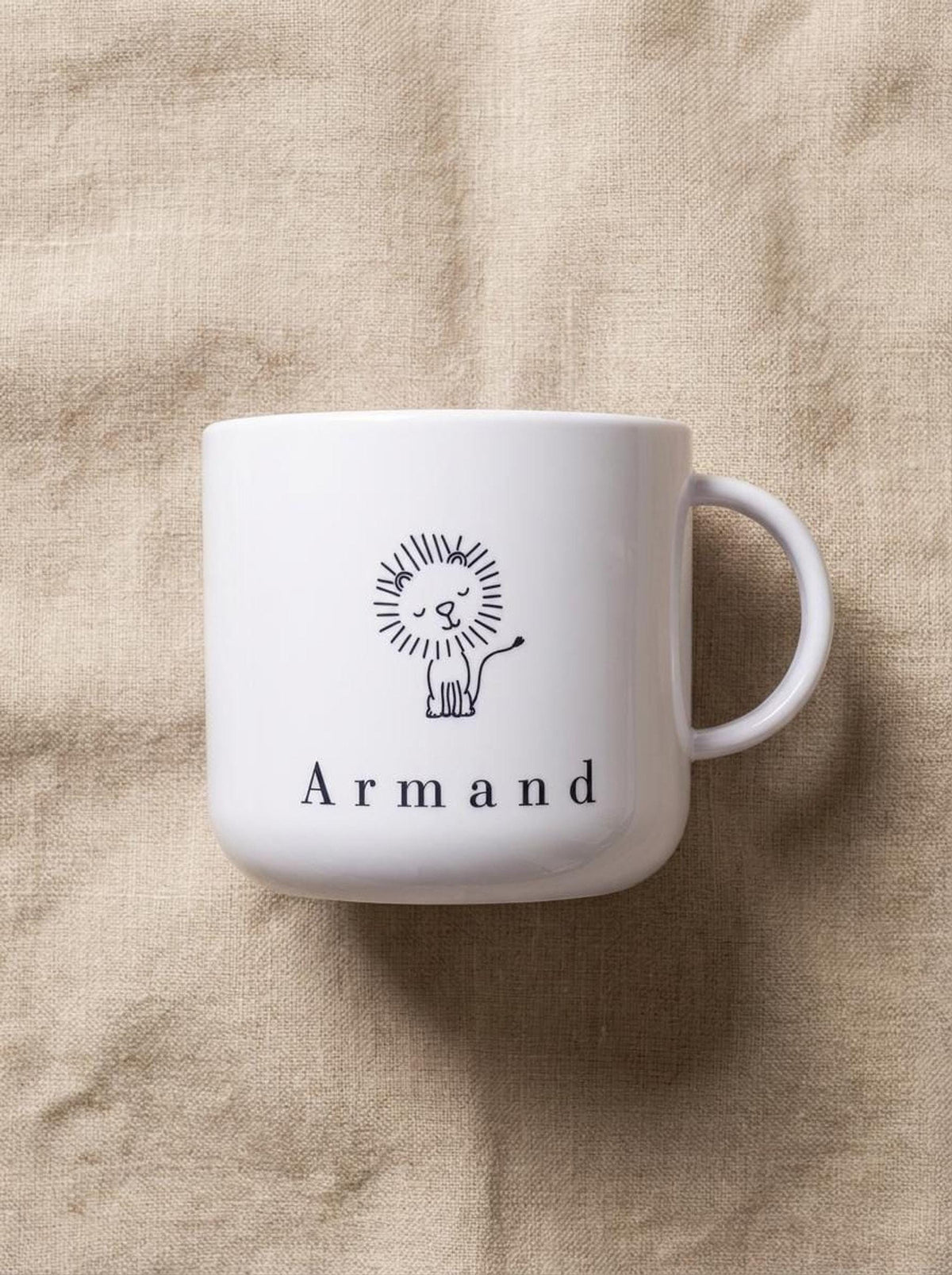 A white polymer cup featuring a line-art lion and the name Armand rests on a neutral-toned linen cloth.