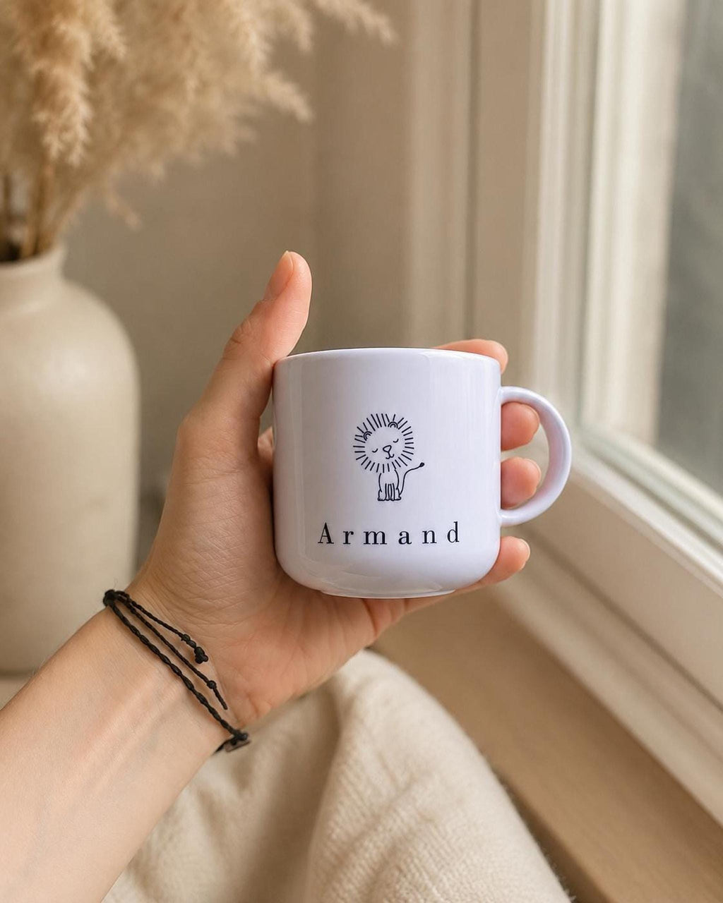 A hand wearing simple bracelets holds the white kids mug near a window with a vase of dried stems in the background.